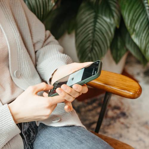 Woman in a beige cardigan sitting in chair looking at phone. Green plan in background.